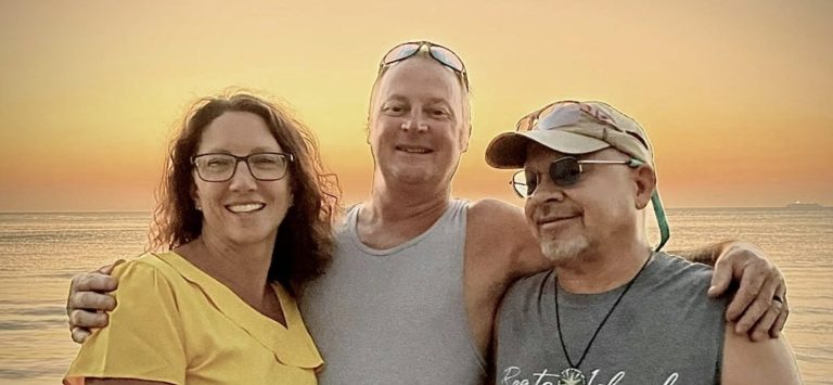 A smiling woman and two men stand close together on the beach at sunset in Roatan, Honduras, with the ocean and a colorful sky in the background.