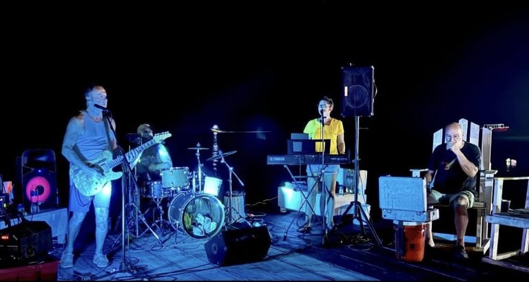 A band performing on a stage at night in Roatan, Honduras, with musicians playing guitar, drums, and keyboard, and the scene illuminated by blue lighting against a dark background.
