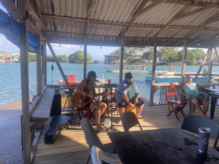 A group of people relax and listen to live music on a wooden dock restaurant by Roatan's water in Honduras, with boats and tropical greenery visible in the background.