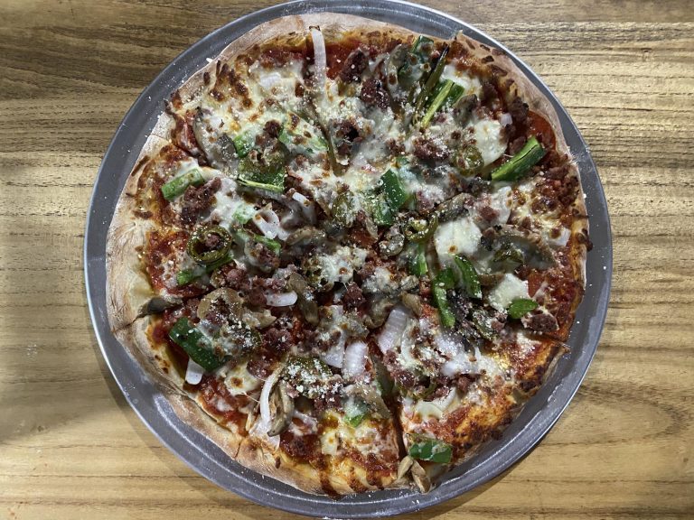 A whole pizza topped with green peppers, onions, ground beef, and melted cheese is displayed on a metal tray on a wooden surface in Roatan, Honduras.