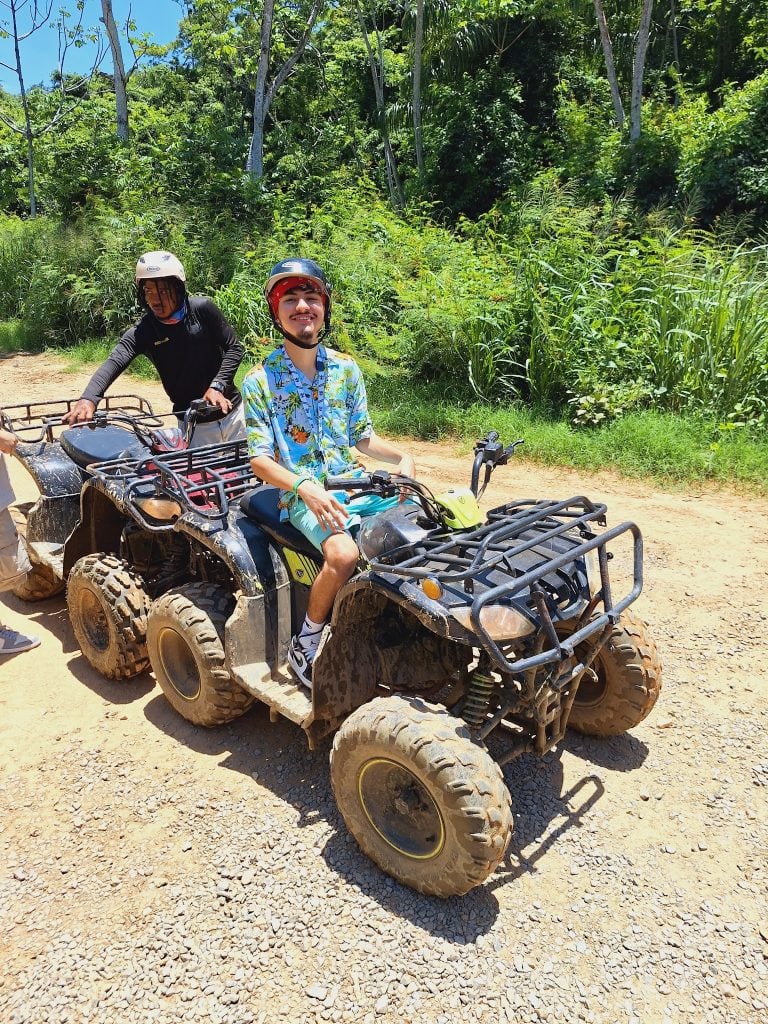 Two people are riding all-terrain vehicles on a dirt path surrounded by lush green tropical vegetation in Roatan, Honduras.