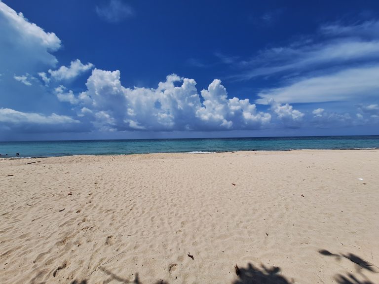A wide sandy beach along Roatan, Honduras, with clear turquoise waters, gentle waves, and a partly cloudy blue sky overhead.