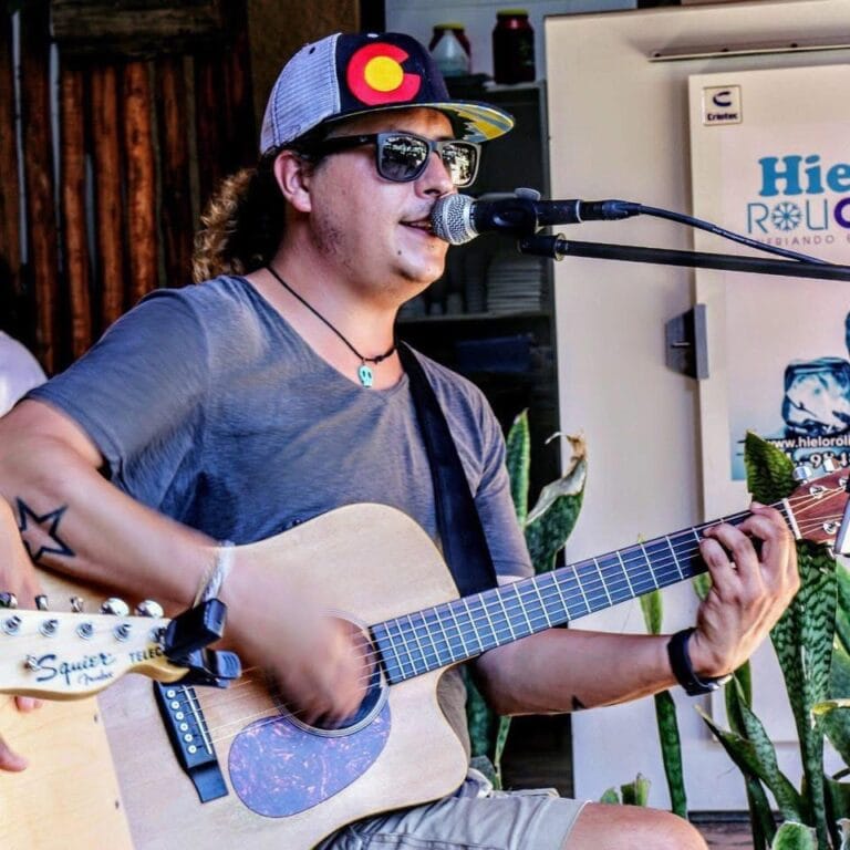 A musician wearing sunglasses and a colorful cap plays an acoustic guitar and sings into a microphone at a live performance in Roatan, Honduras, with potted plants and a promotional banner in the background.