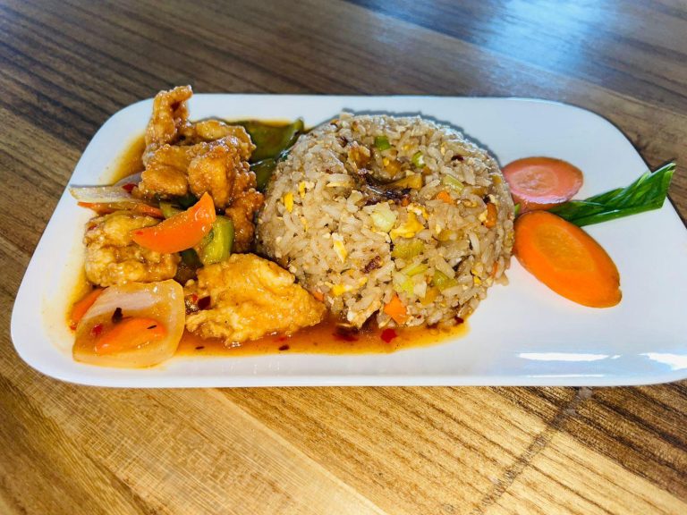 A plate of fried rice with vegetables and scrambled egg, accompanied by sweet and sour chicken with bell peppers and onions, served on a white rectangular dish on a wooden table in Roatan, Honduras.