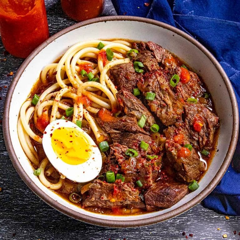 A bowl of Honduran beef stew with noodles, topped with chopped green onions, a halved boiled egg, and red pepper flakes, served on a dark wooden table in Roatan, Honduras.