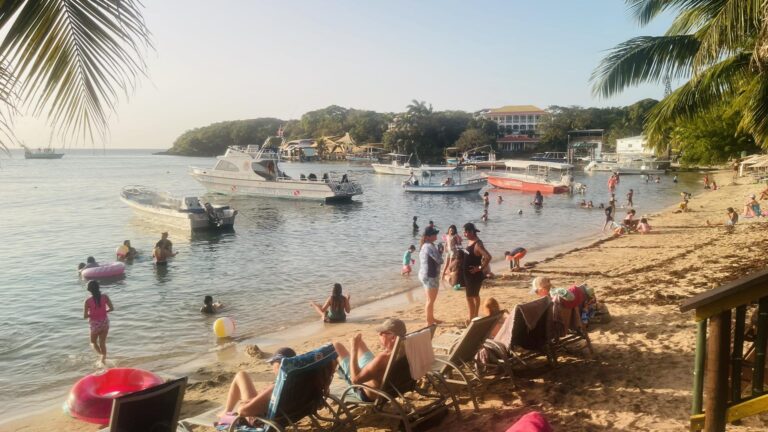 People enjoy swimming and relaxing on the sandy beaches of Roatan, Honduras, with boats anchored near the shoreline and lush trees and buildings in the background.