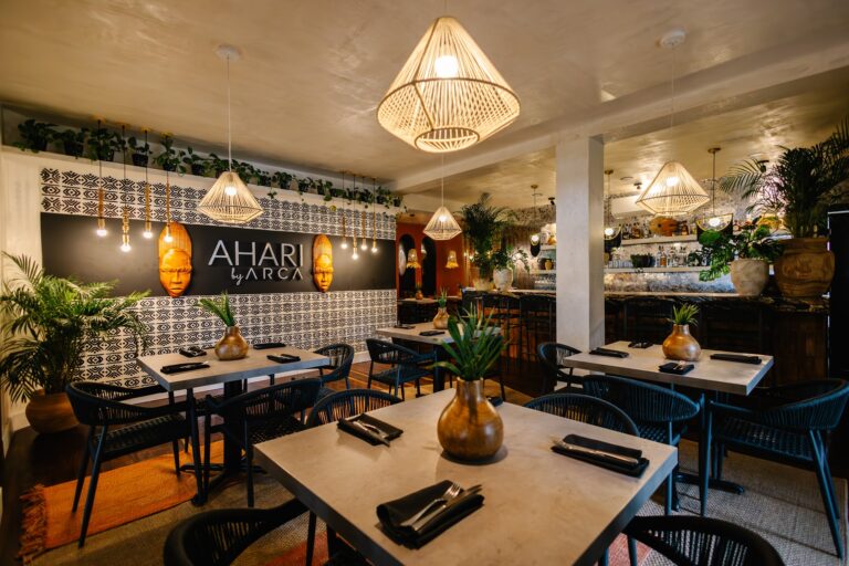 The interior of a stylish restaurant on Roatan in Honduras features white marble tables, black chairs, tropical potted plants, and warm wicker pendant lighting, with decorative masks and greenery enhancing the ambiance.