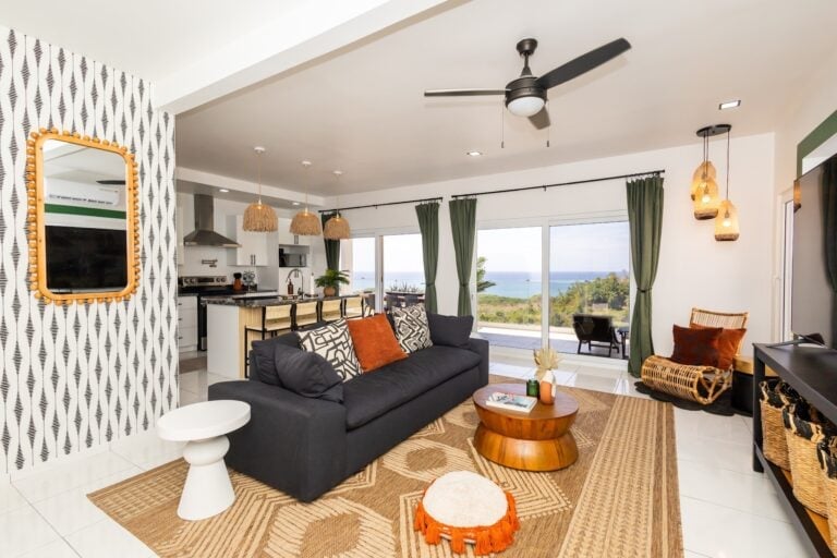 A modern living room in Roatan, Honduras, features a black couch with patterned and rust-colored pillows, large glass sliding doors with green curtains providing ocean views, and decor including woven pendant lights, a mirror with wooden bead frame, and tropical-inspired furniture including a wicker chair and a wooden coffee table.