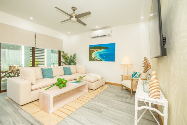 A bright, airy living room in Roatan, Honduras, features a white sofa with turquoise pillows, a wooden coffee table with a decorative green balloon sculpture, a wall-mounted TV, a beach-themed artwork, and a sliding glass door leading to an outdoor patio with wooden furniture.