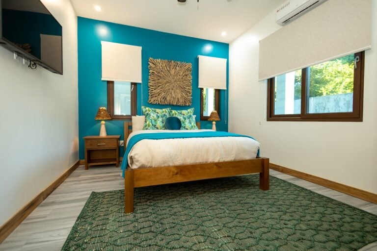 A bedroom in Roatan, Honduras features a wooden bed with tropical-patterned pillows, a teal accent wall with a sunburst wood wall art piece, two windows with beige blinds, wooden nightstands with small lamps, and a television mounted on the wall.