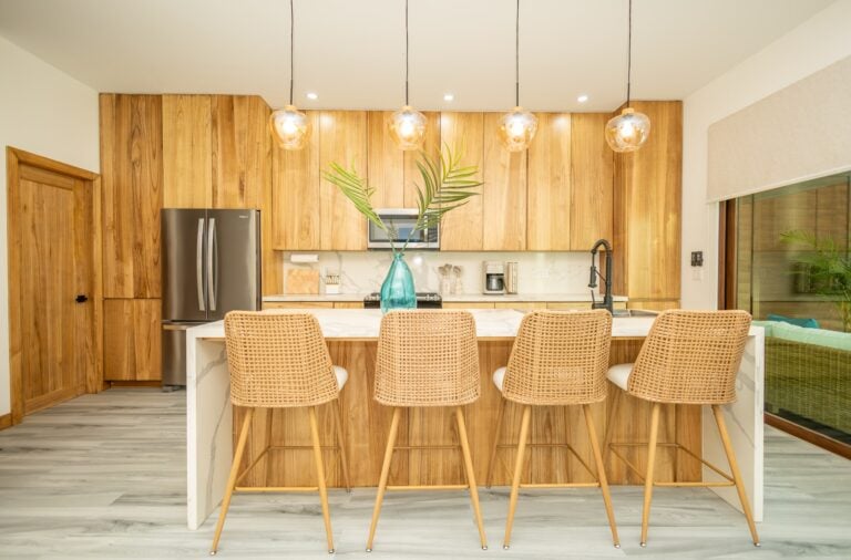 A modern kitchen in Roatan, Honduras features wooden cabinetry, a stainless steel refrigerator, a marble countertop, wicker barstools, and decorative glass pendant lights hanging from a white ceiling.