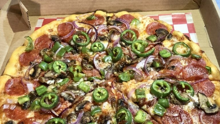A freshly baked pizza topped with pepperoni, green bell peppers, red onions, and mushrooms inside a cardboard box on Roatan, Honduras.
