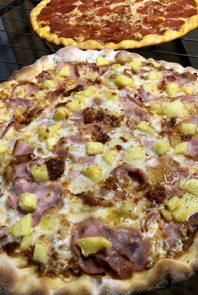 Two freshly baked pizzas topped with pineapple, ham, pepperoni, and bacon are shown cooling on a grill rack, representing a casual dining or food scene in Roatan, Honduras.