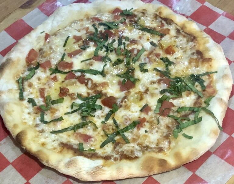 A close-up of a freshly baked pizza topped with cheese, tomato slices, chopped green herbs, and small pieces of meat, served on red and white checkered paper in Roatan, Honduras.