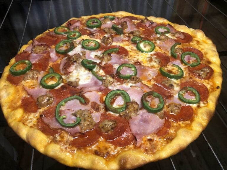 A freshly baked pizza topped with jalapeño slices, ham, sausage, and pepperoni resting on a black oven rack.