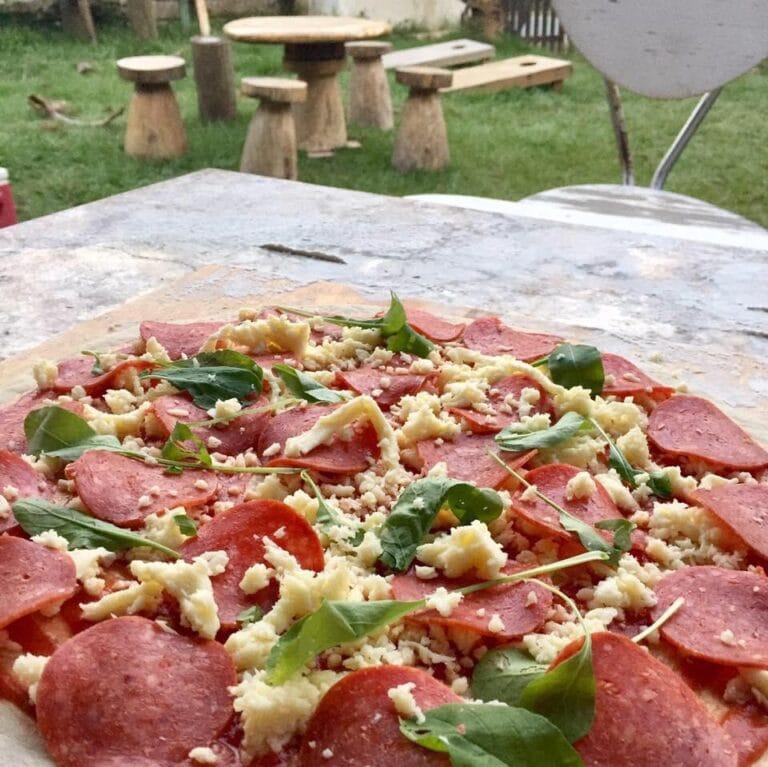 A pepperoni pizza topped with fresh basil leaves and crumbled cheese is placed on a stone table outdoors, with wooden seating and a grassy area in the background in Roatan, Honduras.