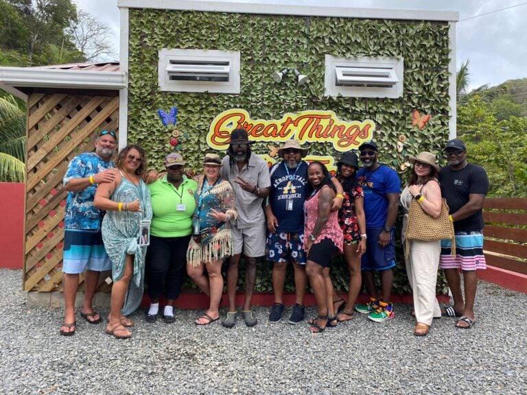 A diverse group of people wearing casual and tropical attire pose in front of a decorative wall with greenery, butterflies, and the sign "Great Things" at a business in Roatan, Honduras.