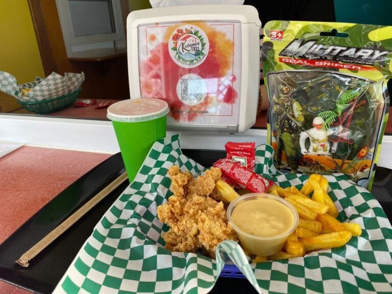 A tray of fried chicken, French fries with dipping sauce, a green paper cup, and a toy soldier figurine in packaging are displayed on a green and white checkered paper liner at a food counter in Roatan, Honduras.