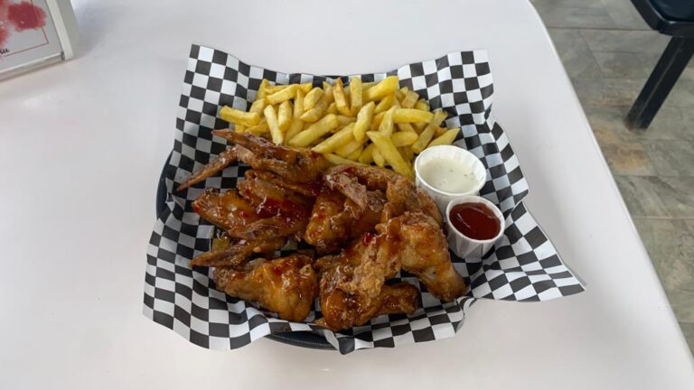 A basket of crispy chicken wings topped with sauce, alongside a portion of French fries and two small cups of dipping sauces, placed on a white table at a restaurant in Roatan, Honduras.
