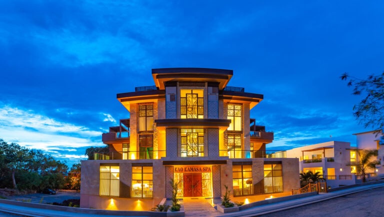 A modern three-story spa building with illuminated interior lighting and stained glass windows, located on a hillside in Roatan, Honduras, during dusk with a partly cloudy sky overhead.