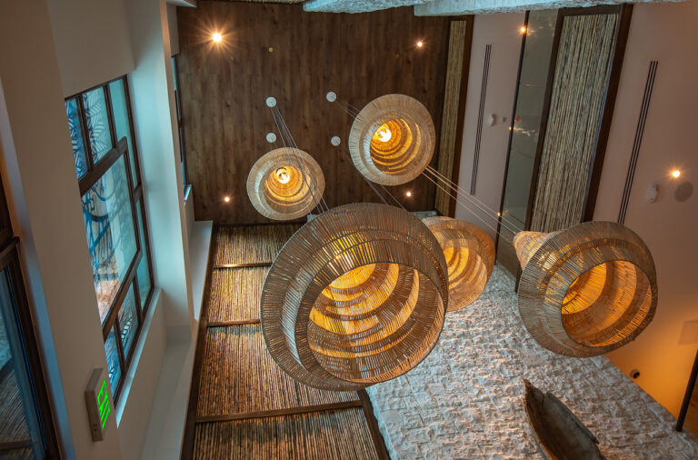 A view of an indoor space in Roatan, Honduras, showcasing large, woven, circular pendant lights hanging from the ceiling over a textured stone floor and wooden wall, with large windows on the side allowing natural light to enter.
