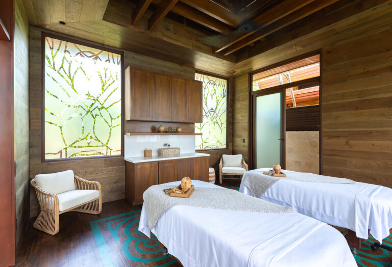 A spa room in Roatan, Honduras features two massage beds with rolled towels and wooden massage tools, set against wooden walls with large decorative windows and natural light.