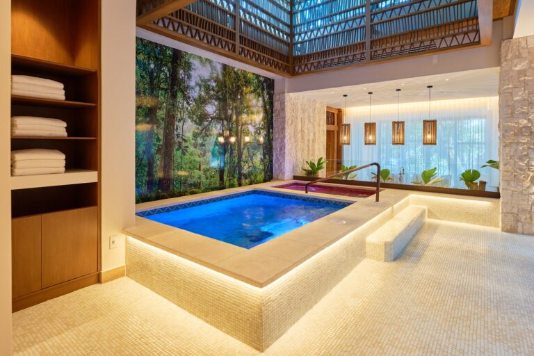 A luxurious indoor swimming pool area featuring a small spa, surrounded by beige mosaic tiles, natural stone walls, and large windows with sheer curtains, in a resort located in Roatan, Honduras.