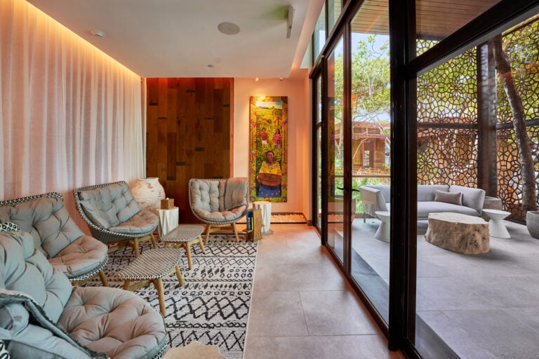The interior of a modern, brightly lit lounge area featuring beige tufted chairs, a black-and-white patterned rug, colorful artwork depicting Honduran culture, and large glass doors opening to a shaded outdoor patio with seating and decorative wooden screens in Roatan, Honduras.