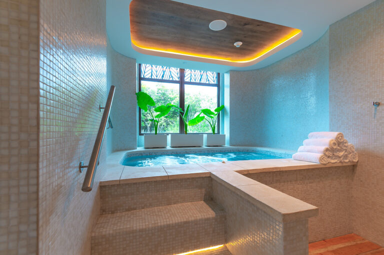A luxurious indoor jacuzzi in a spa room on Roatan, Honduras, featuring beige mosaic tile walls, a wooden ceiling accent with embedded lighting, three rolled white towels, and large windows with potted tropical plants overlooking greenery.