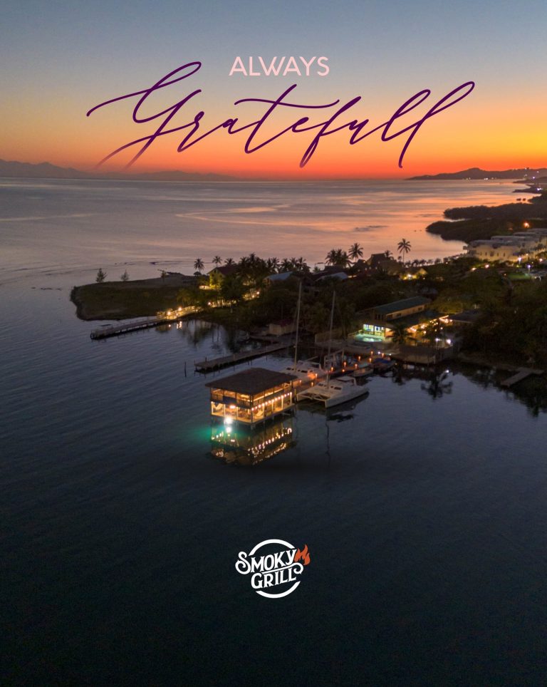 Aerial view of a marina in Roatan, Honduras during sunset with illuminated boats and waterfront buildings, featuring the Smoky Grill logo at the bottom.