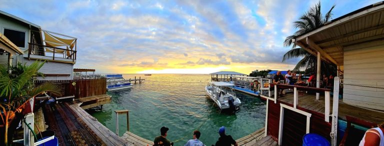 A waterfront dining area on Roatan, Honduras, features a wooden deck with people enjoying drinks and sunset views over the Caribbean Sea, surrounded by boats docked at piers and lush palm trees.