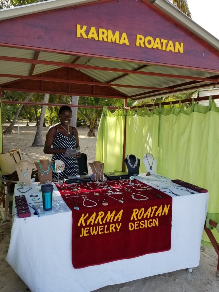 A woman sells handmade jewelry at a beachside stall labeled "Karma Roatan Jewelry Design" on Roatan, Honduras, with tropical palm trees and sandy beach visible in the background.