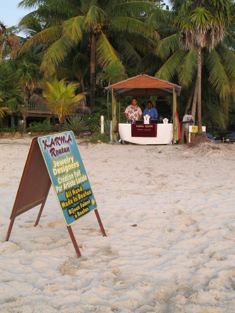 A small booth under a wooden canopy on the sandy beach of Roatan, Honduras, displays jewelry created by local designers, with two people standing behind the table and palm trees in the background. A colorful sign in the foreground advertises jewelry handmade in Roatan.