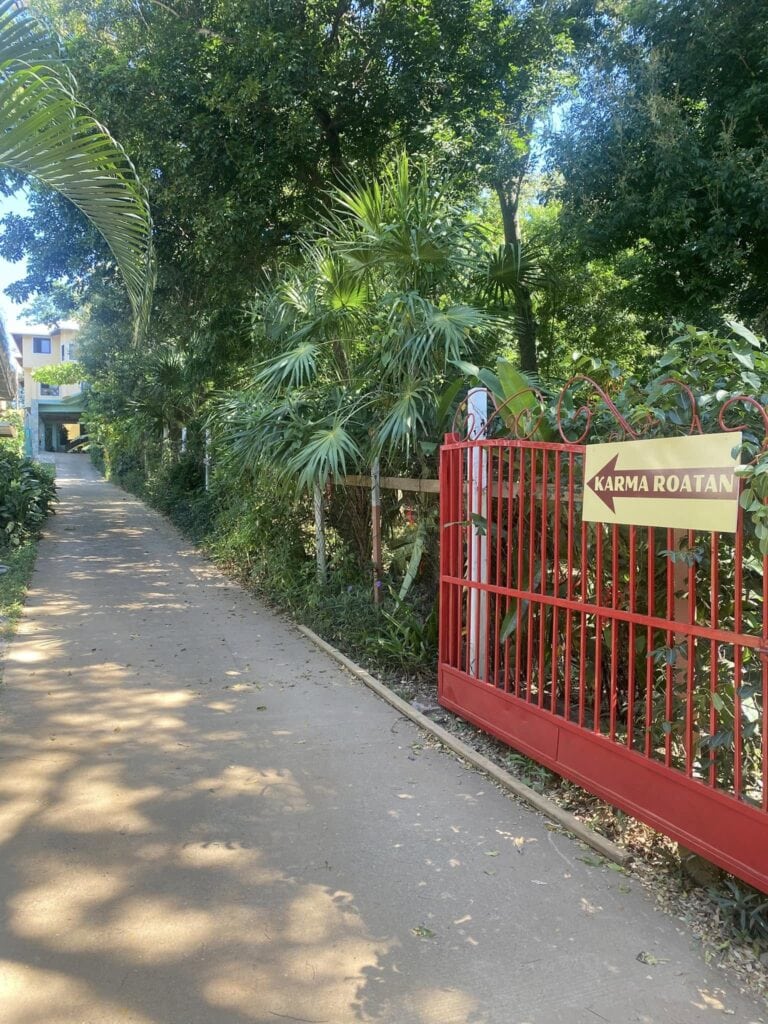 A red gate with a sign pointing left that reads "Karma Roatan" is located along a narrow concrete pathway surrounded by lush green tropical foliage in Roatan, Honduras.