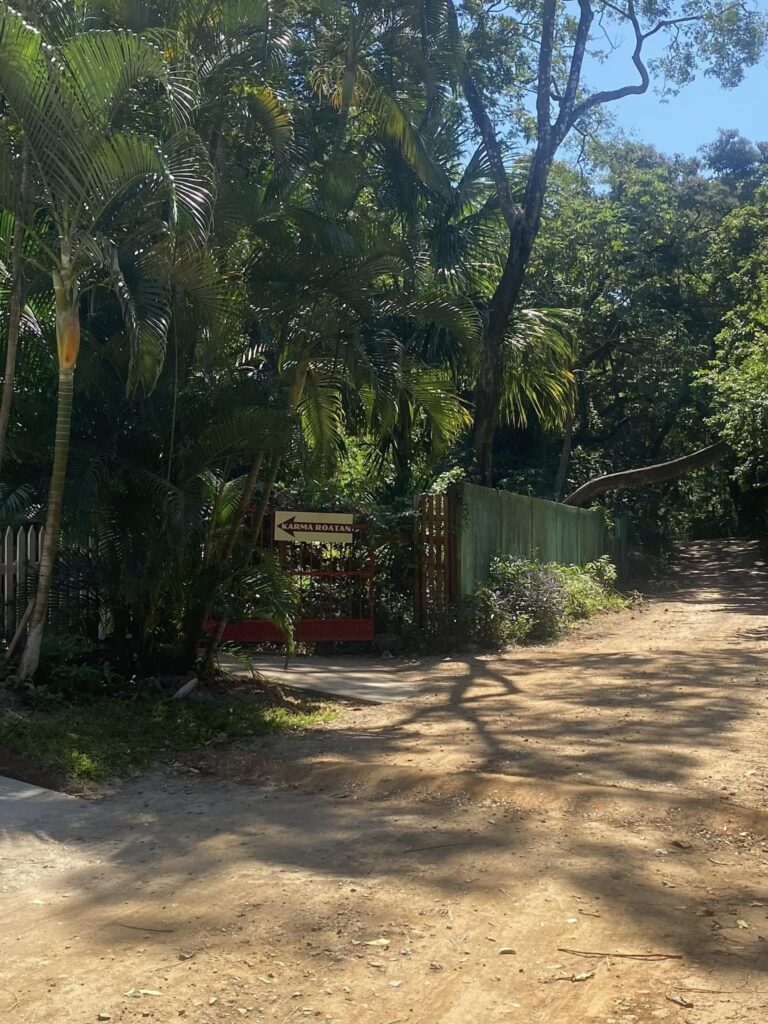 A dirt pathway lined with lush green trees and foliage leads to a gate with a sign that reads "Karma Roatan" in Roatan, Honduras.