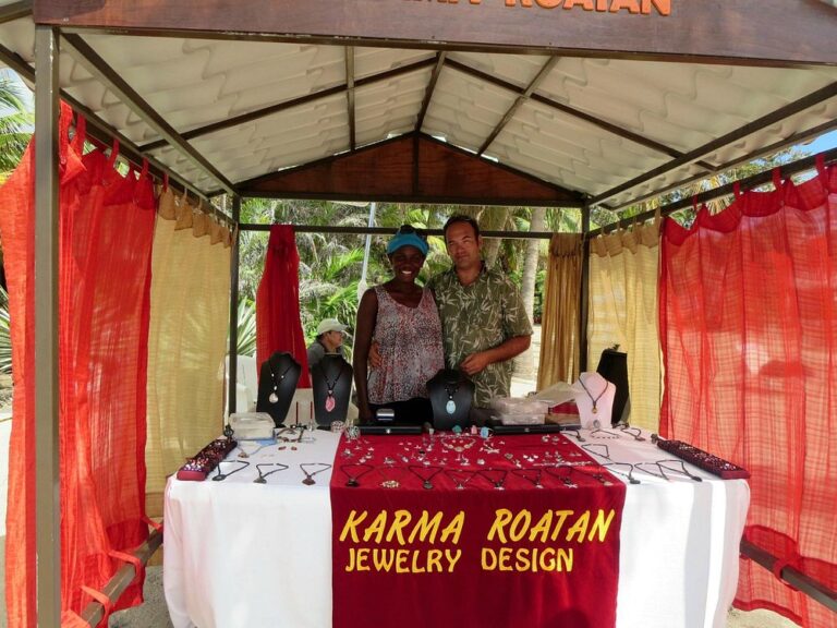 A jewelry booth displaying handcrafted necklaces, bracelets, and pendants under a shaded outdoor canopy at a market in Roatan, Honduras, with two vendors standing behind the table and colorful fabric curtains on the sides.