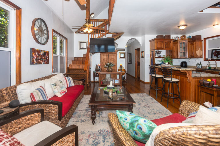 Cozy living room in a Honduran home featuring wicker furniture, a red cushioned sofa, tropical-themed pillows, and a view of the wooden staircase in Roatan, Honduras.
