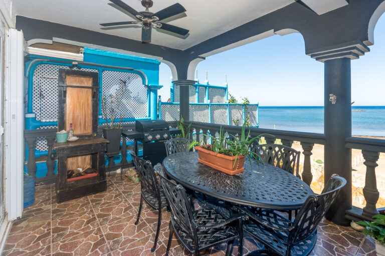 Seaside balcony in Roatan, Honduras, featuring a round metal dining table with six chairs, potted succulents, and a view of the ocean through decorative balustrades.