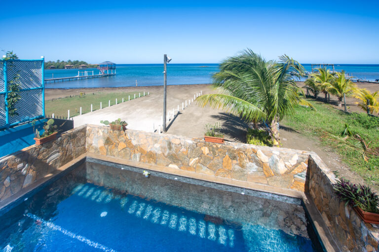View from a hillside terrace over a swimming pool towards a sandy beach with palm trees on Roatan, Honduras, with the Caribbean Sea in the background.