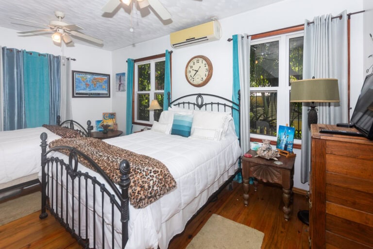 Cozy bedroom with white walls, wooden floors, and tropical decor, featuring a metal bed frame, multiple windows with blue and white curtains, and decorations highlighting Roatan and Honduras.