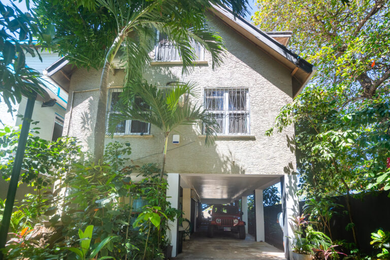 Two-story house in Roatan, Honduras, with a textured beige exterior, protected windows with decorative metal bars, and surrounded by lush tropical plants including palm trees and flowering shrubs.