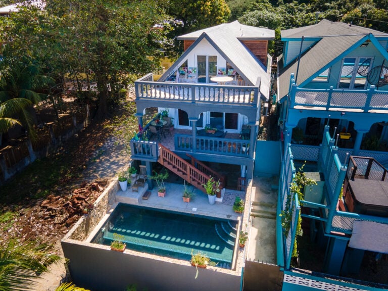 Three-story white house with multiple balconies and a swimming pool in Roatan, Honduras, surrounded by lush trees and neighboring homes on the island paradise.