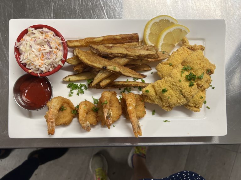 A plate of fried shrimp, potato wedges, coleslaw, lemon slices, and dipping sauces served on a white rectangular dish in a restaurant on Roatan, Honduras.