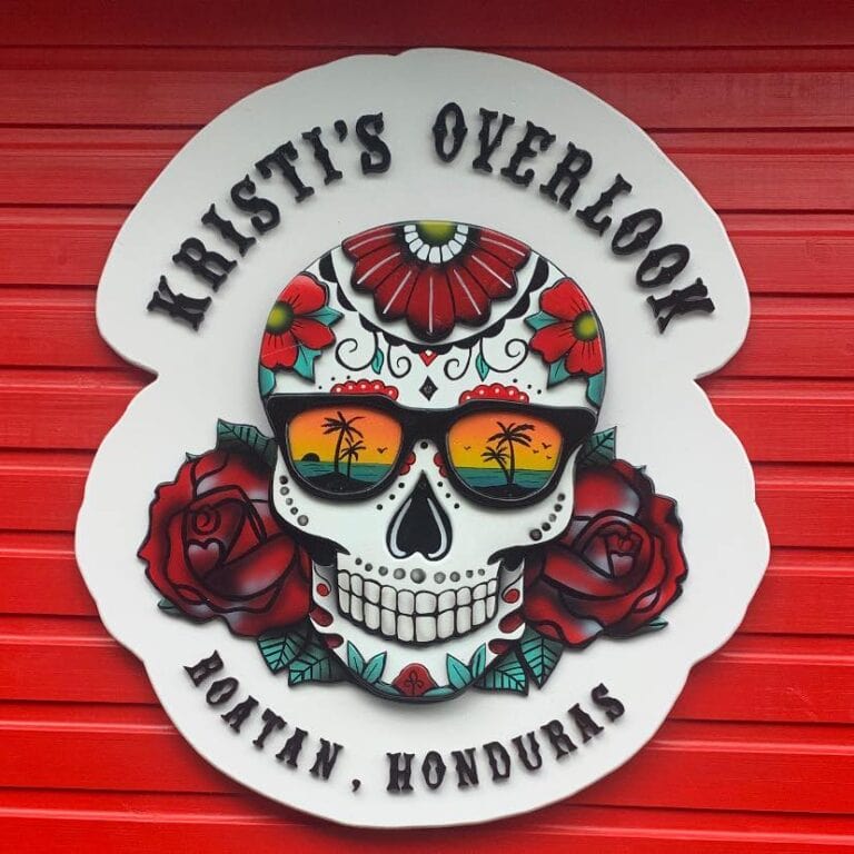 A colorful sugar skull with floral designs and sunglasses reflecting a sunset with palm trees, mounted on a white background with the phrase "Kirsti's Overlook" and "Roatan, Honduras" written above and below, against a red wooden wall.