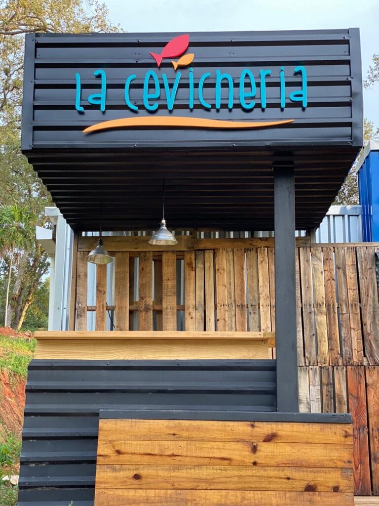 A newly constructed wooden and metal food stand with a sign reading "la cevicheria" in blue letters, located in Roatan, Honduras, featuring hanging pendant lights and a partially enclosed serving area.