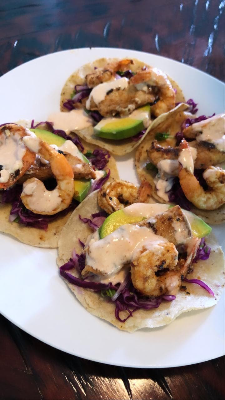 Four shrimp tacos topped with grilled shrimp, avocado slices, shredded purple cabbage, and drizzled with a creamy sauce are served on a white plate, set against a dark wooden table in Roatan, Honduras.