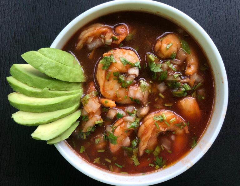 A bowl of shrimp ceviche garnished with chopped onions, cilantro, and lime slices on the side, served with a dark sauce, displayed on a black textured surface in Roatan, Honduras.