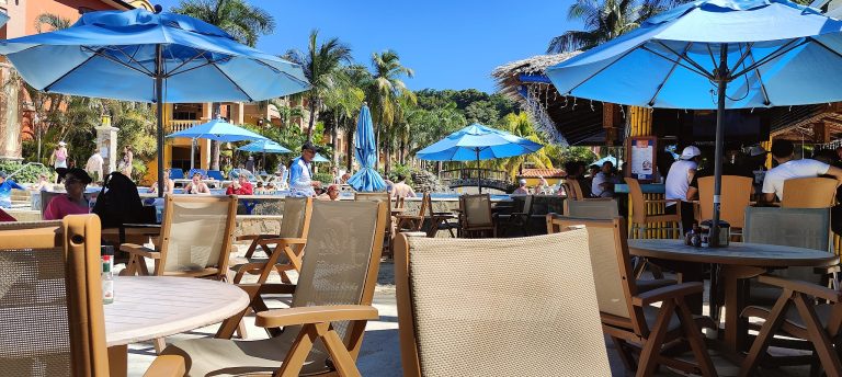 An outdoor dining area at a restaurant in Roatan, Honduras, featuring wooden tables and chairs, large blue umbrellas, and people relaxing by the pool with tropical palm trees and lush greenery in the background.