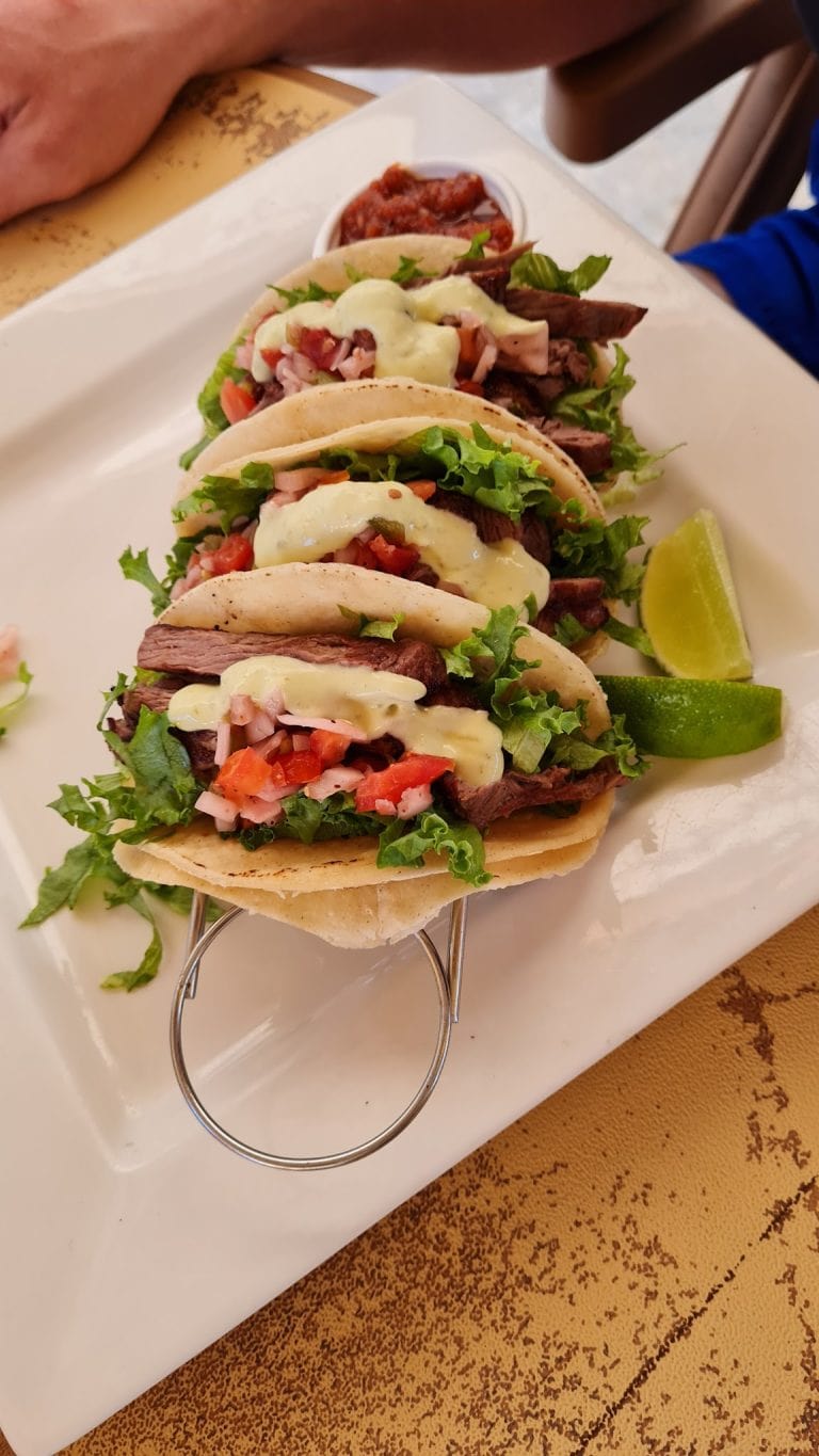 Three beef tacos with lettuce, chopped tomatoes, melted cheese, and cilantro served with lime wedges, accompanied by a side of salsa, on a white rectangular plate in Roatan, Honduras.