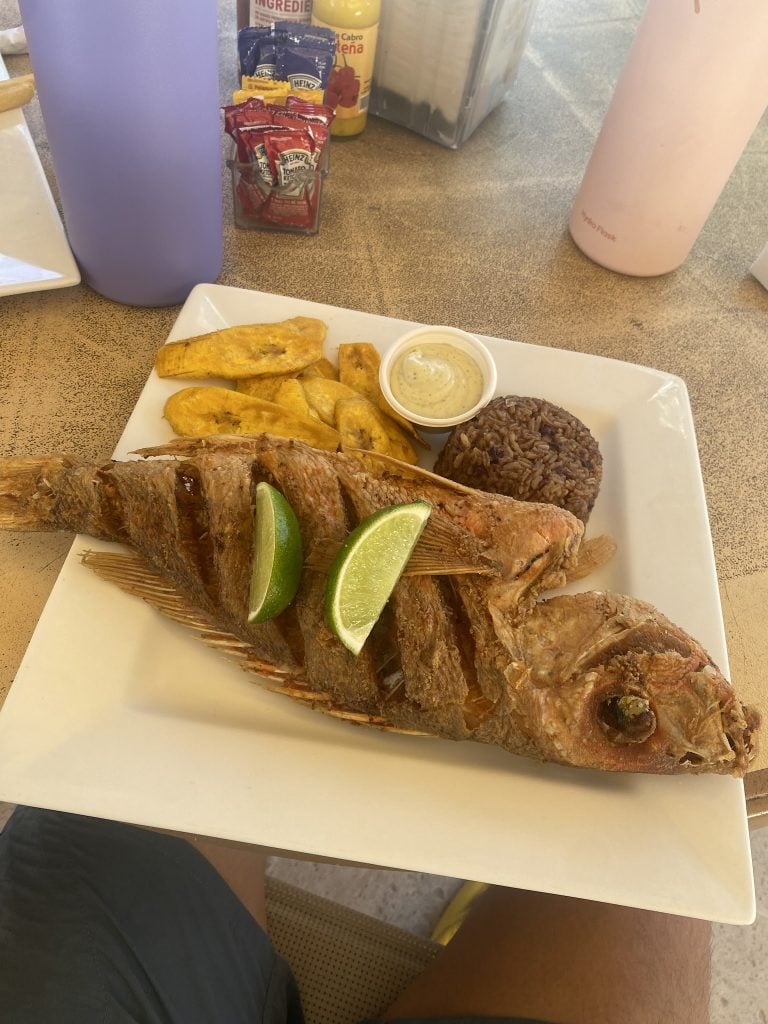 A whole cooked fish served with lime wedges, fried plantains, rice and beans, and a side of dipping sauce on a white plate at a restaurant in Roatan, Honduras.